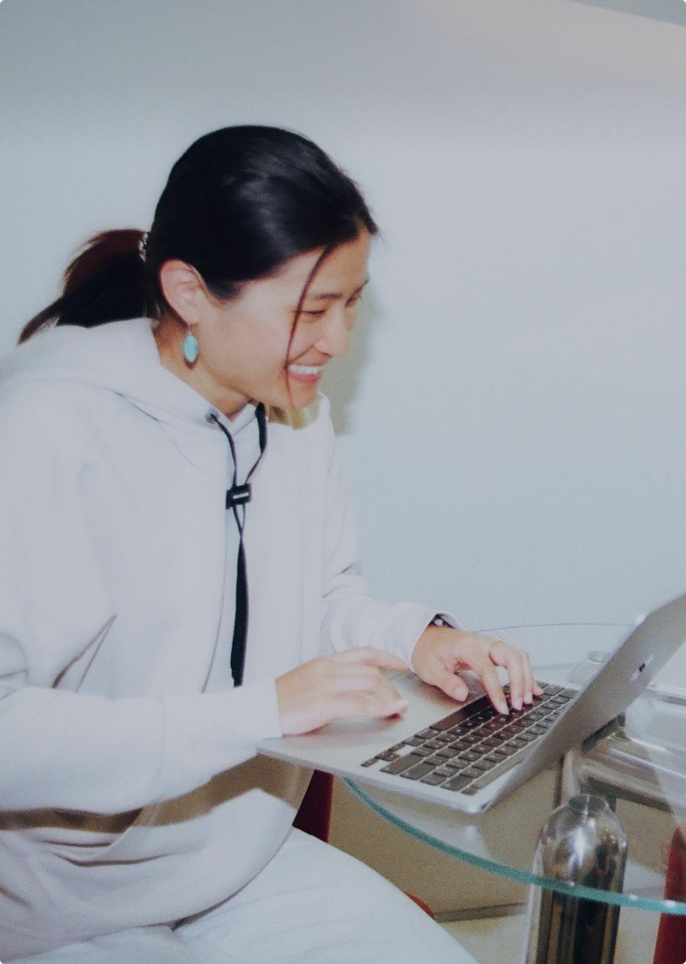 Smiling woman with her hair tied back wearing a light hoodie and turquoise earrings types on a laptop at a glass table.