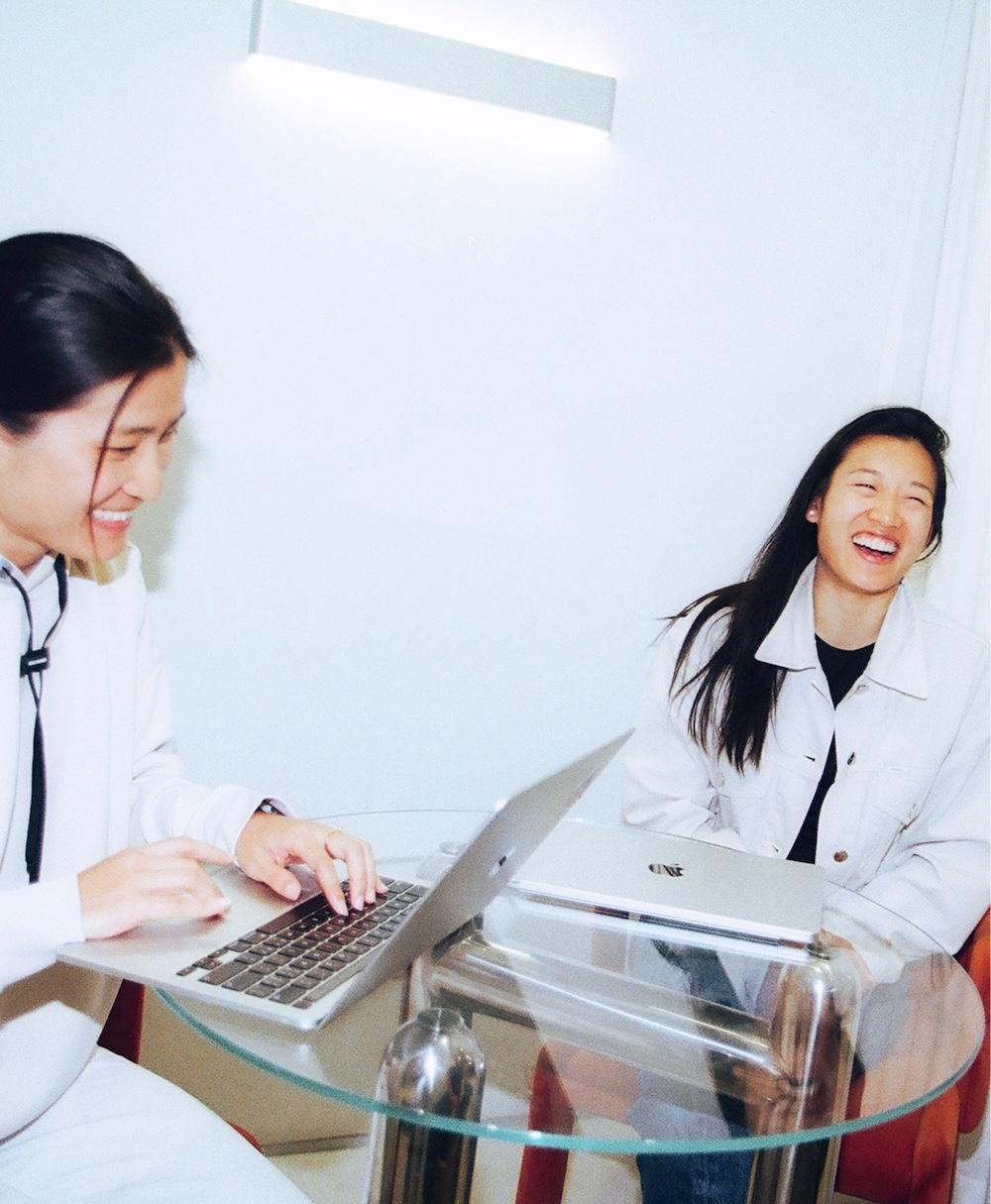 Two people sit at a round glass table with laptops, one typing and the other smiling.