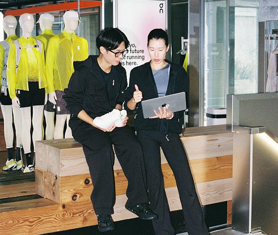 Two people in black athletic clothing sit on a wooden bench in a store, one holding a white sneaker while the other gestures at a laptop, with mannequins in neon yellow jackets displayed behind them.