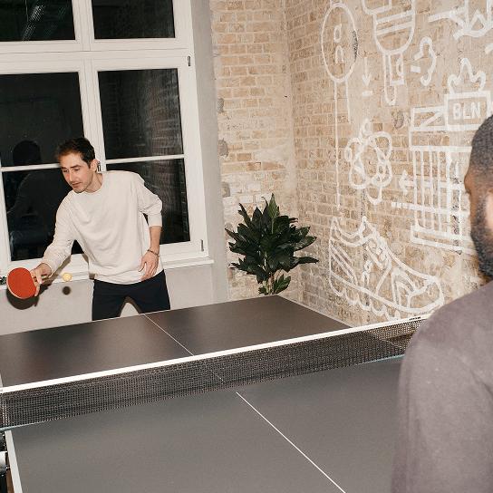 Two people play table tennis indoors, one hitting the ball with a red paddle while a partially visible opponent stands at the other end and a brick wall with a mural fills the background.