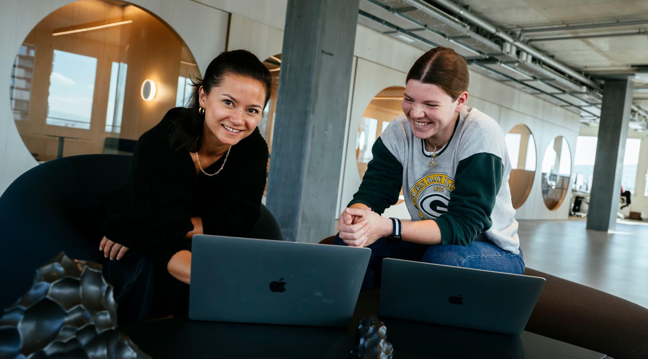 Two people sat at laptops whilst smiling, one is looking at the camera.