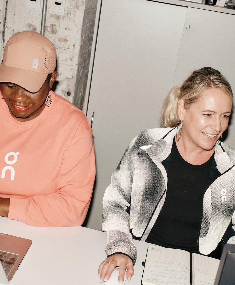 Two women sit side by side at a desk, smiling and working with a laptop and an open notebook.