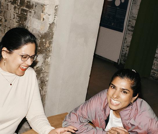 Two people seated at a wooden table against a brick wall, smiling and leaning toward each other.