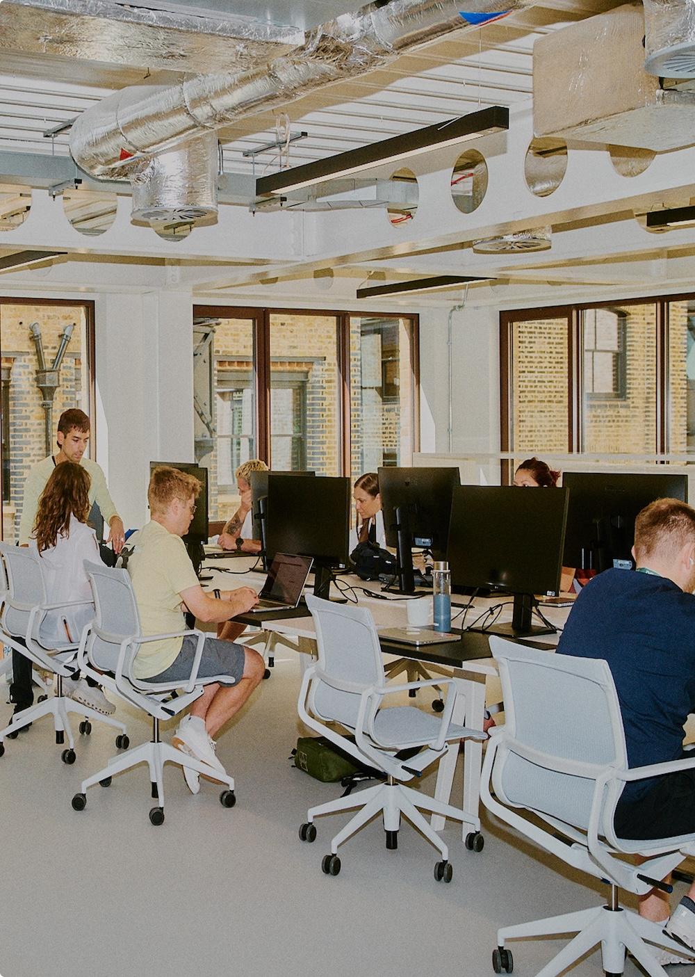 Open-plan office with people seated at white rolling chairs working on laptops and desktop monitors at long desks beneath exposed ductwork and large windows.