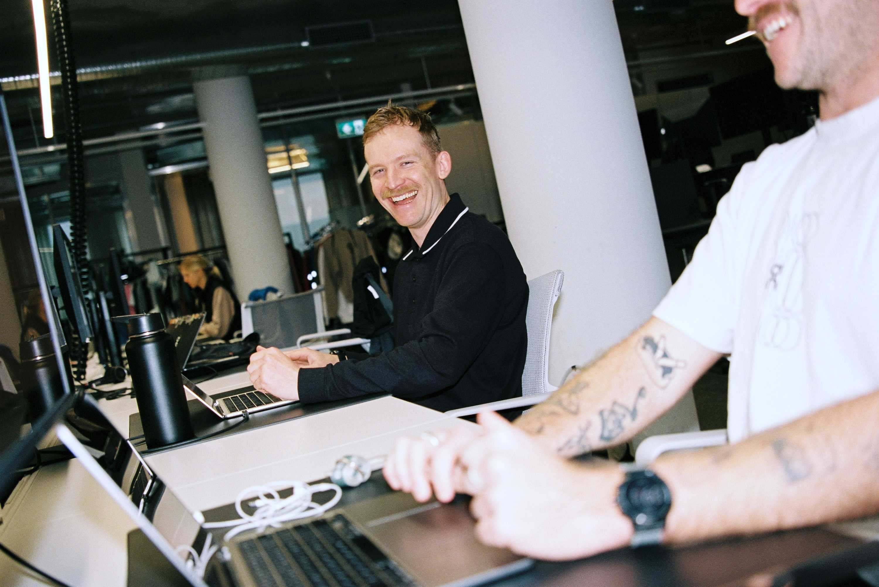 A smiling man in a black shirt sits at a desk using a laptop in an open office while a tattooed coworker in a white shirt is visible in the foreground.