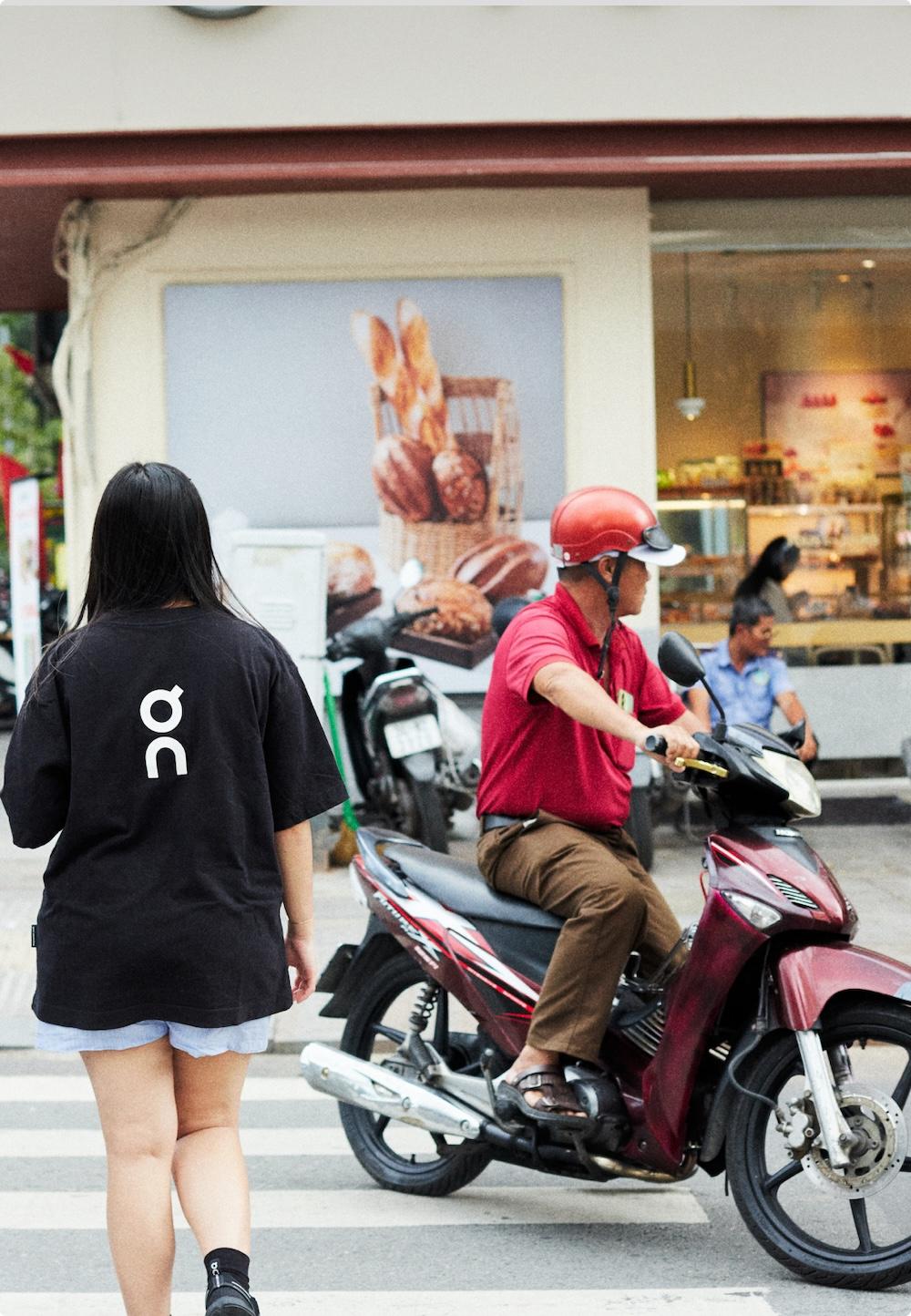 A woman in a black T-shirt walks across a crosswalk past a man wearing a red helmet seated on a red scooter in front of a bakery storefront displaying bread.