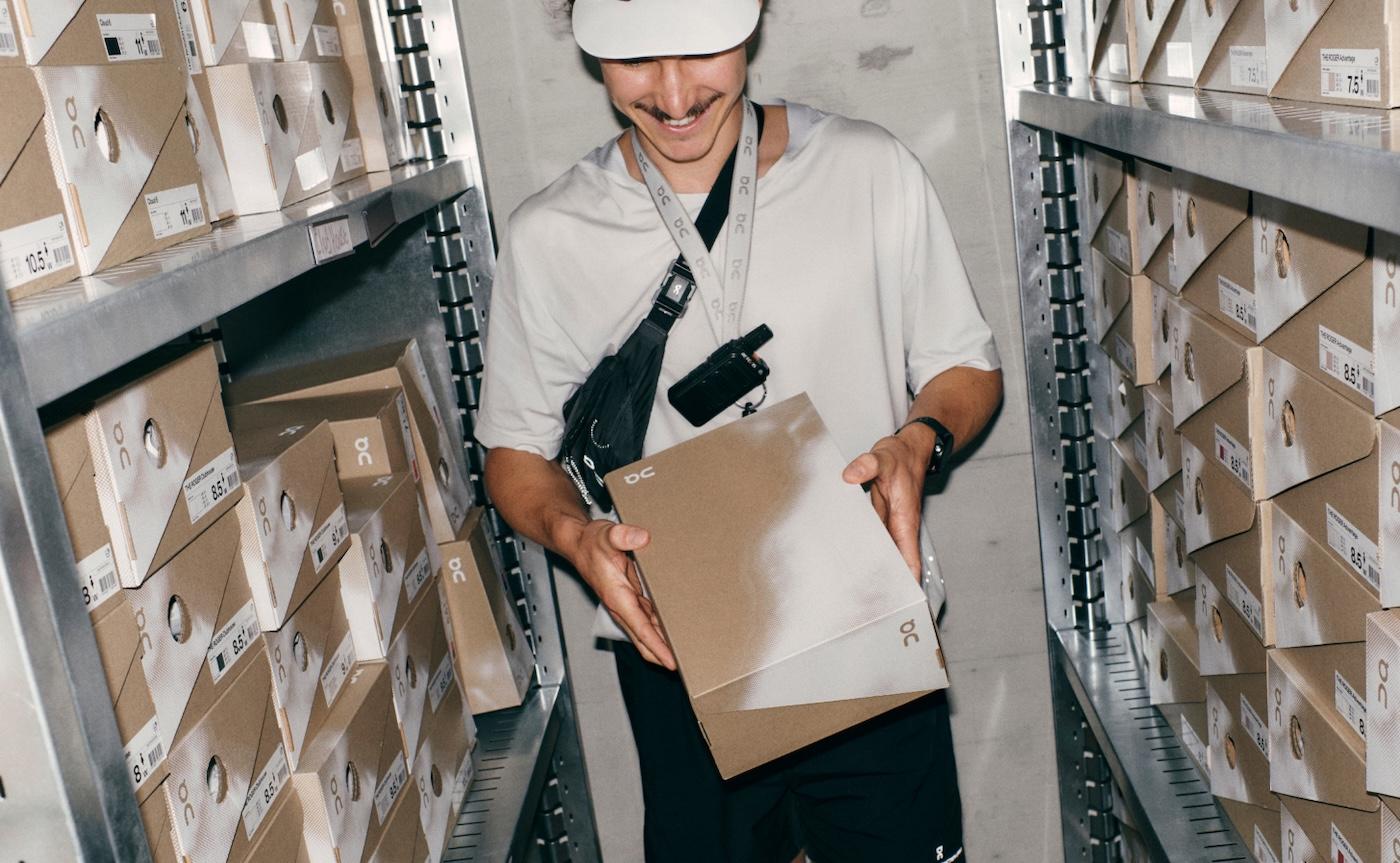A smiling person in a white cap and t-shirt looks down while holding a brown shoebox in front of metal shelving stocked with similar boxes.