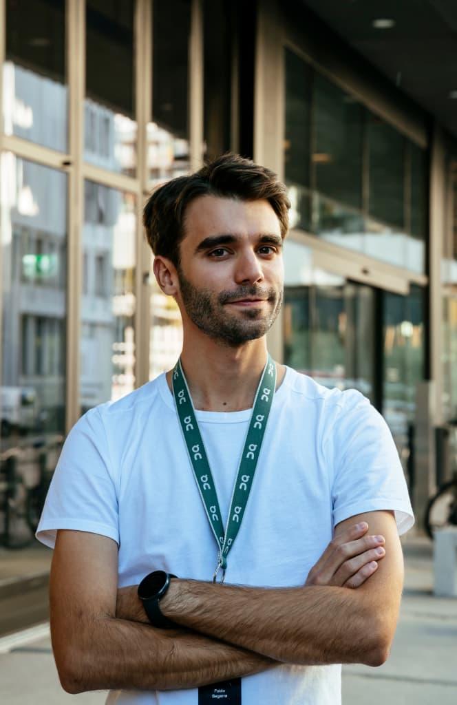 Man standing in a white T-Shirt with an On lanyard with his arms crossed and smiling at the camera
