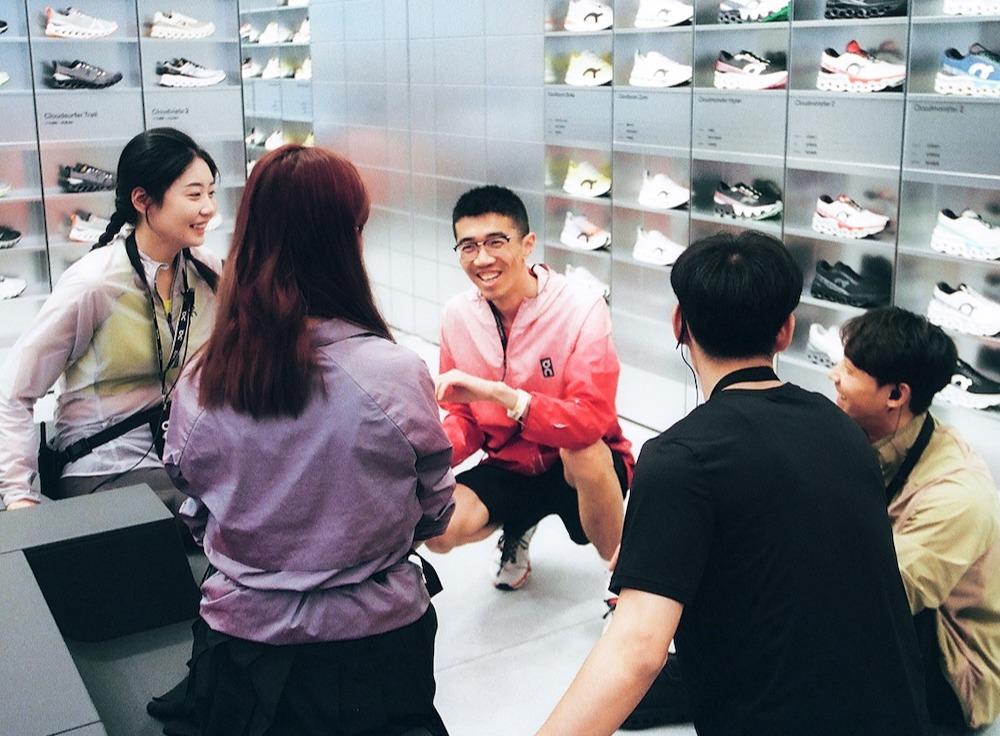 Group of five store employees in sporty attire sit and chat, smiling, in front of a wall display of sneakers.
