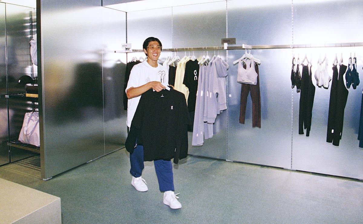 A smiling person holds up a black sweatshirt in front of a clothing rack displaying hoodies and activewear in a mirrored retail space.