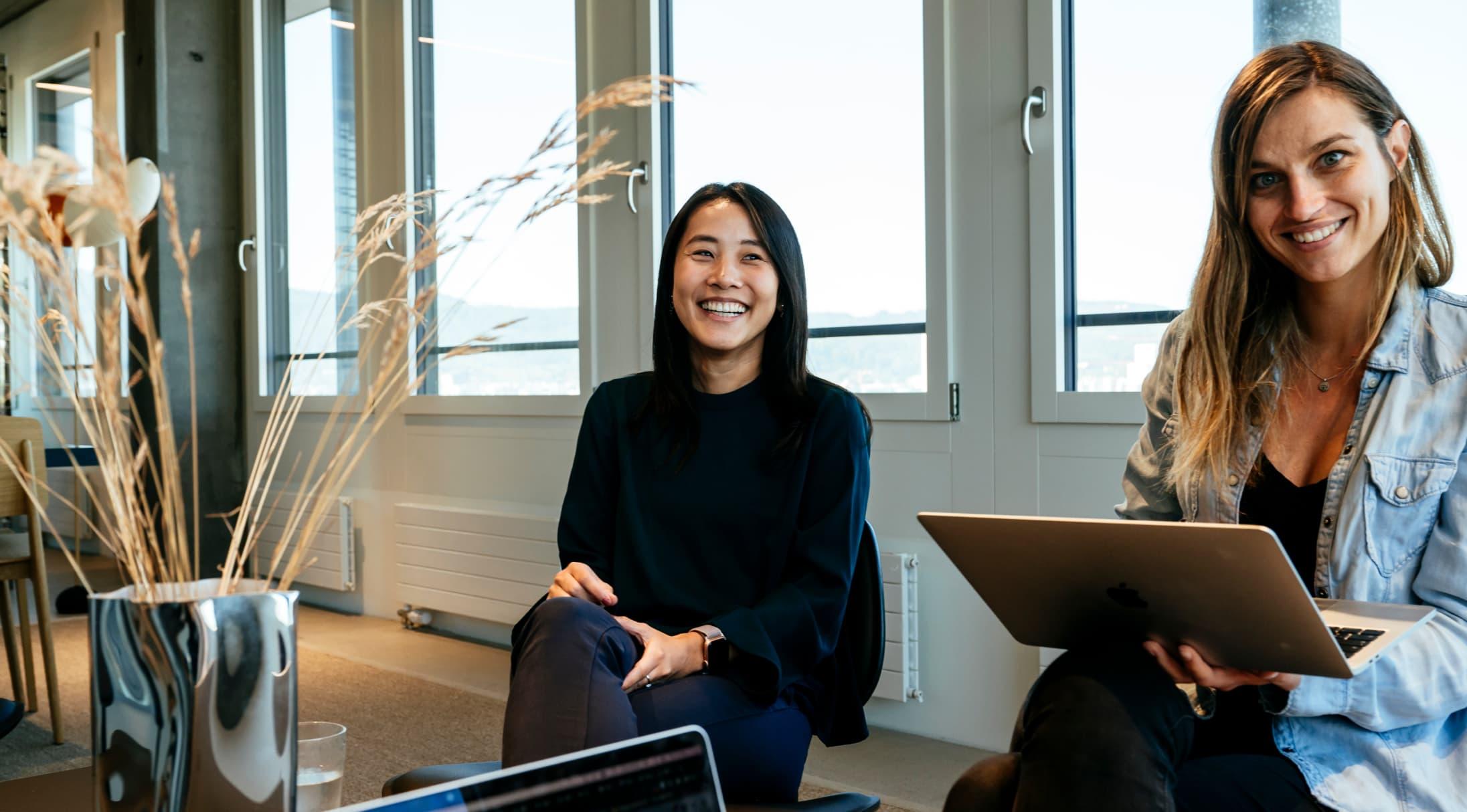 Two women sat with laptops, smiling at the camera.