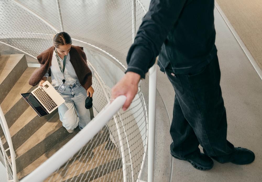 A person walks up a curved staircase carrying an open laptop and headphones while another person stands above holding the railing.