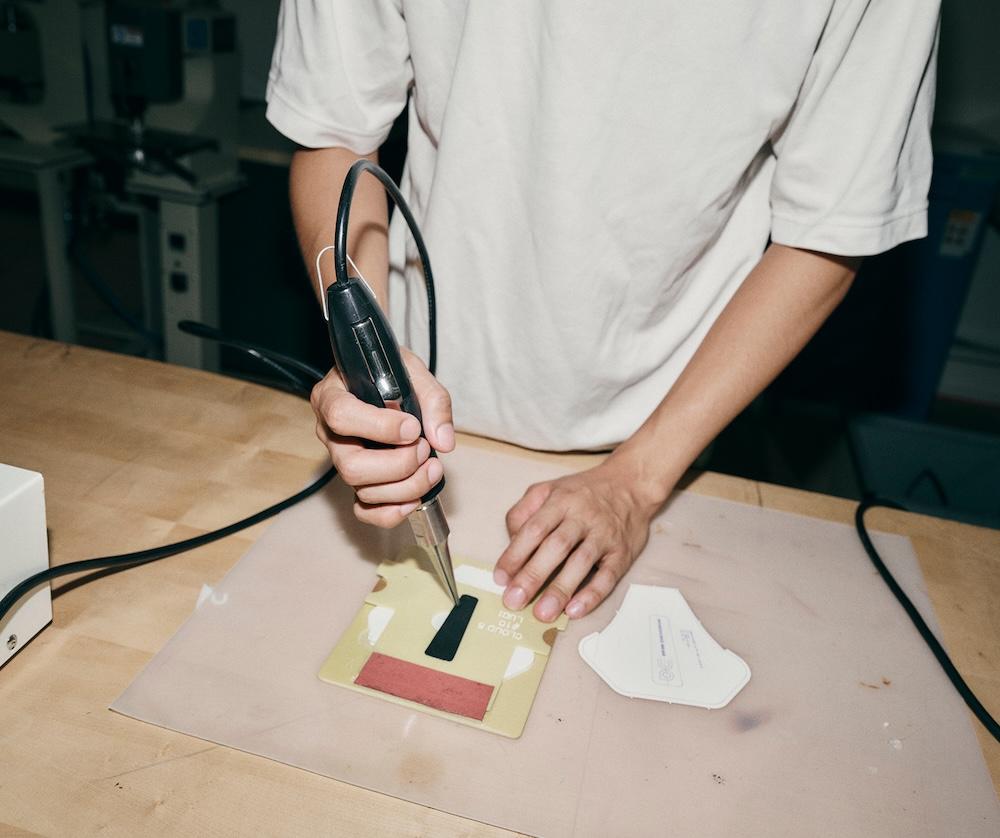 A person holds a soldering iron over a small yellow card with a black rectangular piece on a workbench.