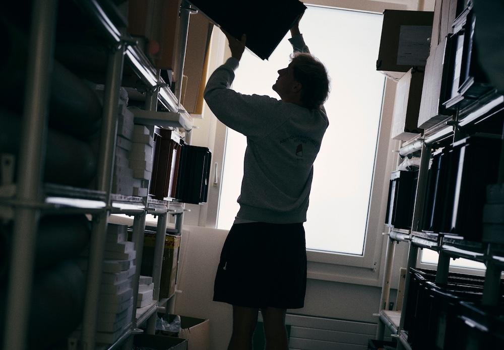 Person standing between metal shelving units in a storage room reaches up to a box on an upper shelf near a bright window.