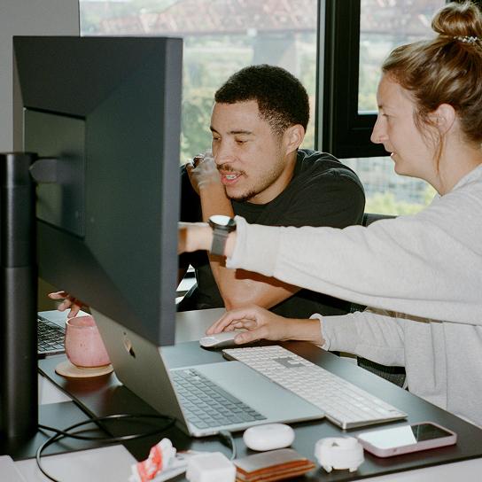 Two people collaborate at a desk, with one person pointing at a large monitor while the other uses a mouse beside a laptop.
