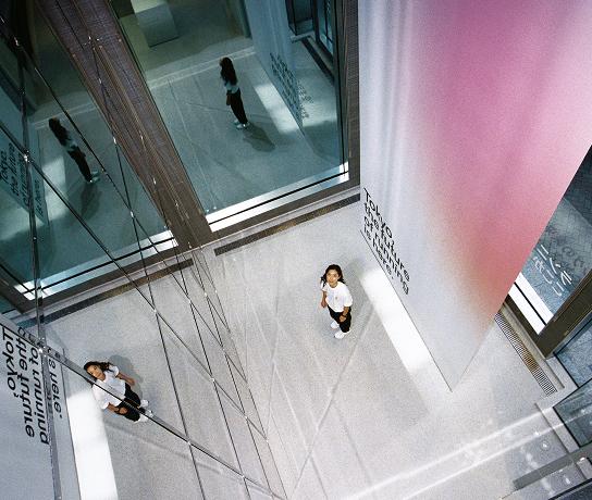 Two individuals stand in a multi-level glass atrium seen from above, with reflections on the glass walls and a tall wall panel showing a pink-to-white gradient.