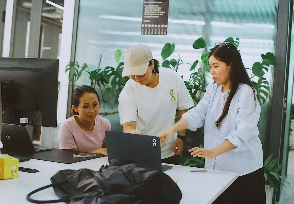 Three people gathered around a laptop at a table in an office with potted plants, with one person pointing at the laptop screen as the others look on.
