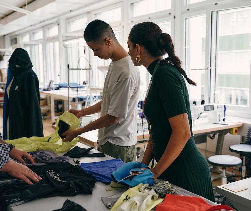 Two people stand at a worktable in a sewing workshop, examining colorful fabric pieces and garments with sewing machines and materials in the background.