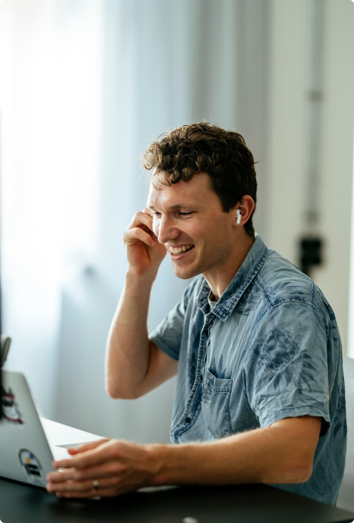 A man sitting at his laptop is listening to something through his earphones and smiling