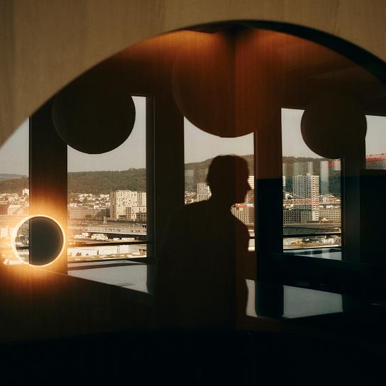 Silhouetted person reflected in an interior window with round light fixtures, overlooking a cityscape of buildings and distant hills.