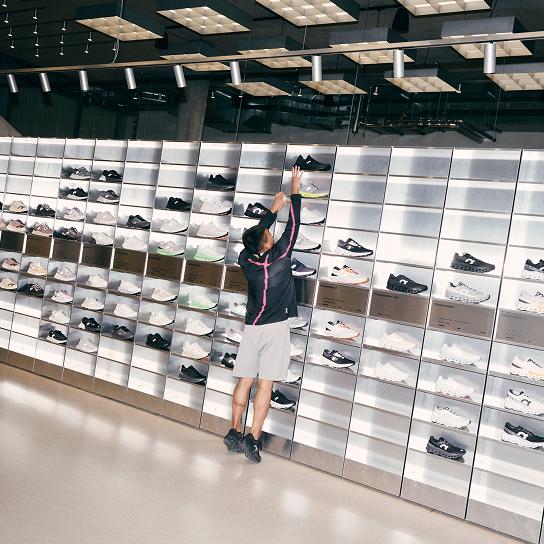 A person reaching up toward a sneaker on a high shelf in a wall display of athletic shoes.