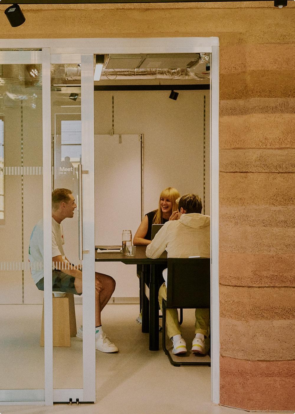 Three people sit around a table in a glass-walled meeting room with a laptop and water bottle, talking and smiling.