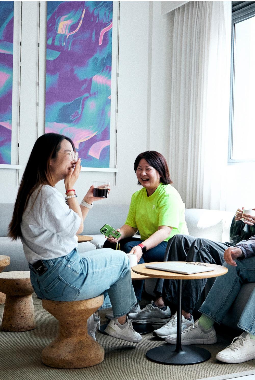 Three people sit in a bright lounge area talking and laughing over beverages, one holding a phone, with a closed laptop on a small table and colorful abstract artwork on the wall.