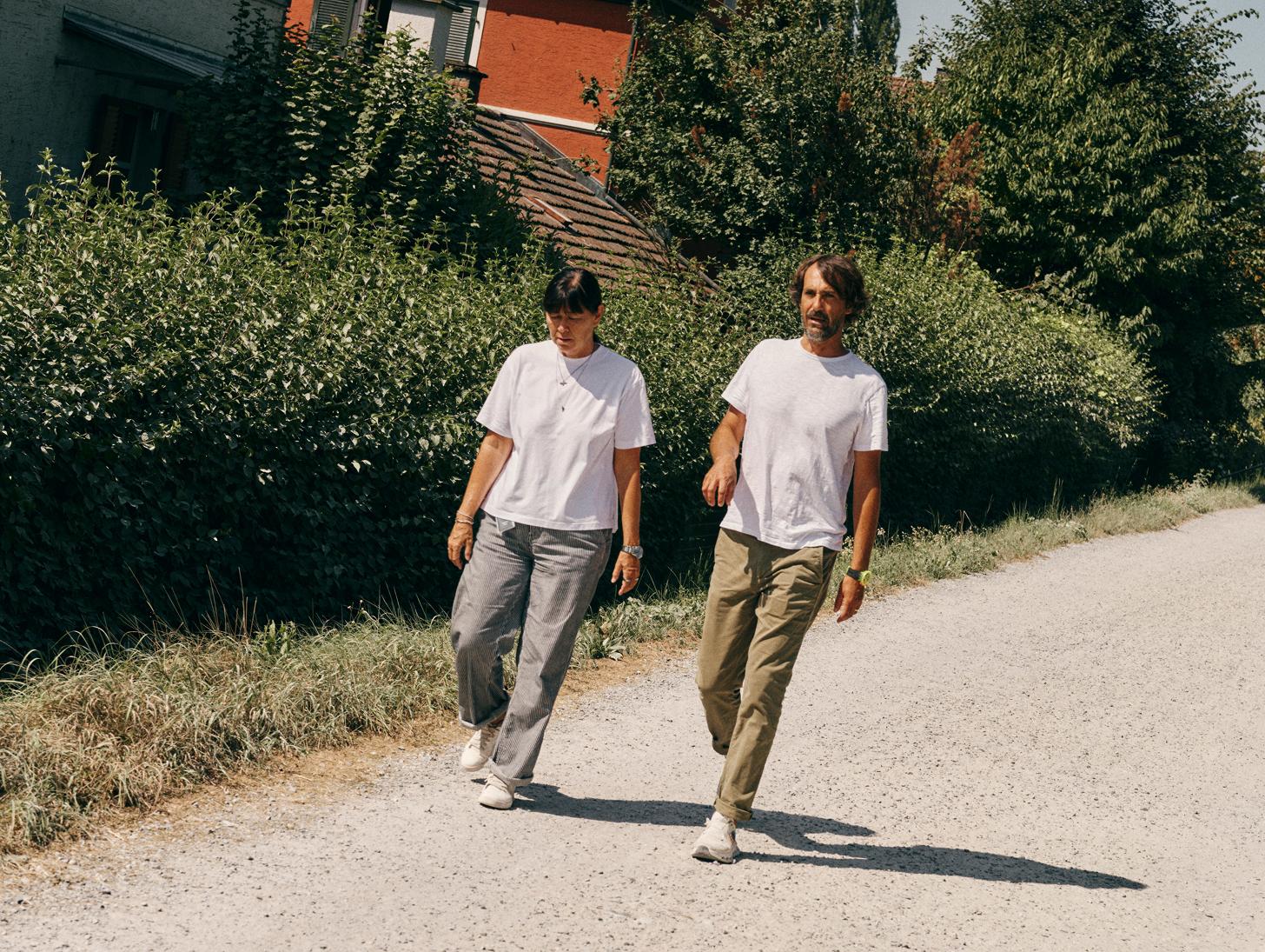 A man and a woman wearing white T-shirts walk side by side on a sunlit gravel path beside dense hedges and a house.