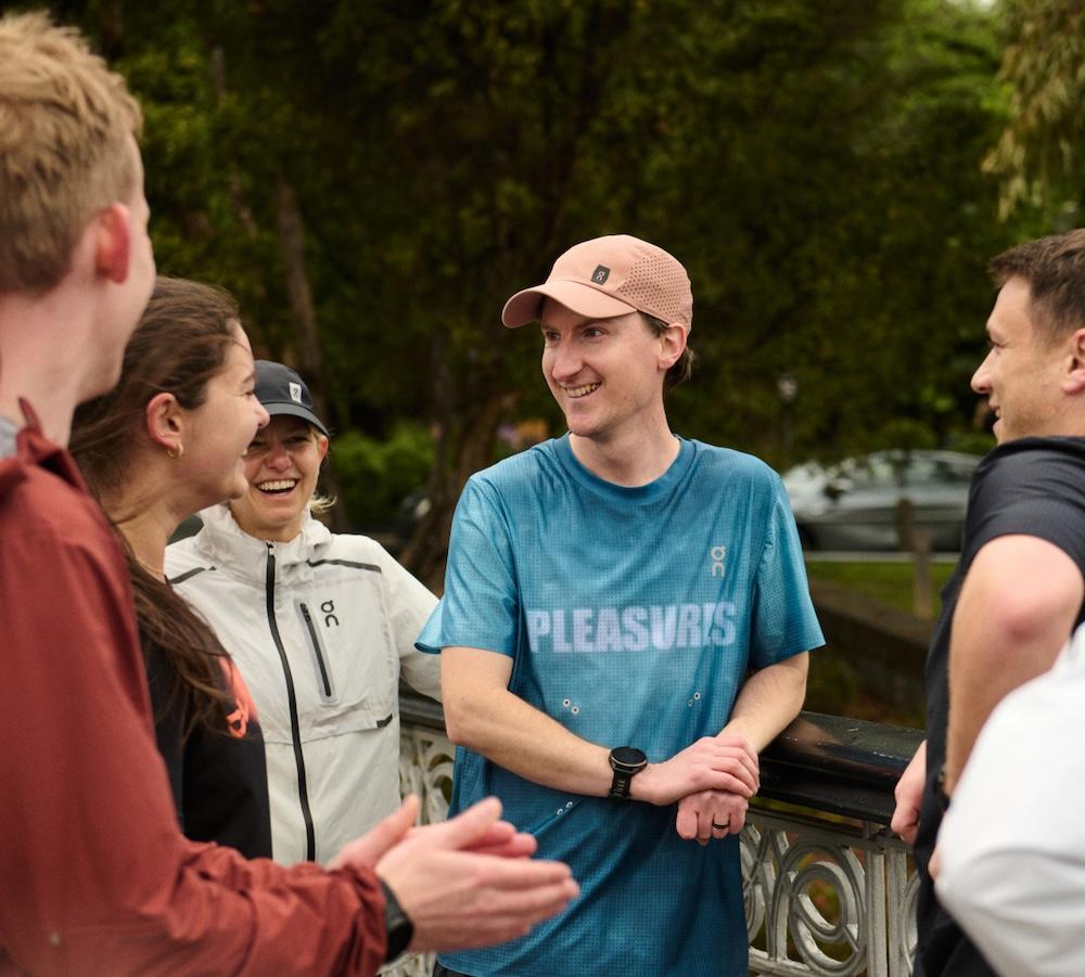 Several adults stand outdoors by a decorative railing, smiling and talking as a man in a blue shirt and pink cap leans on the railing.