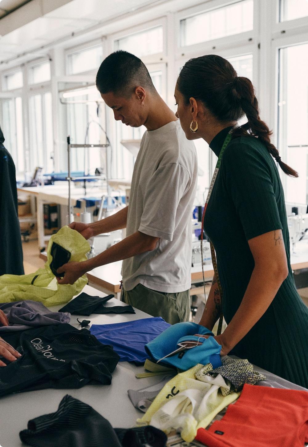 Two people examine colorful fabric samples on a table in a bright design studio with sewing machines.