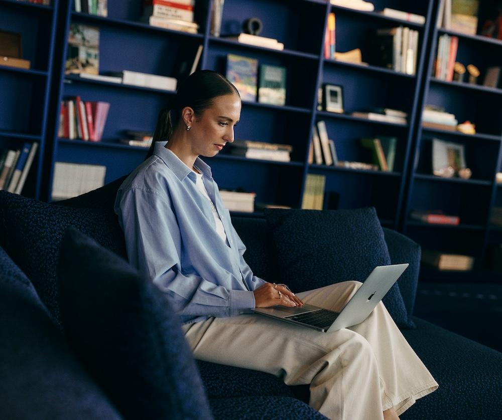 A woman sits on a dark blue sofa typing on a laptop with a blue bookshelf filled with books and objects behind her.