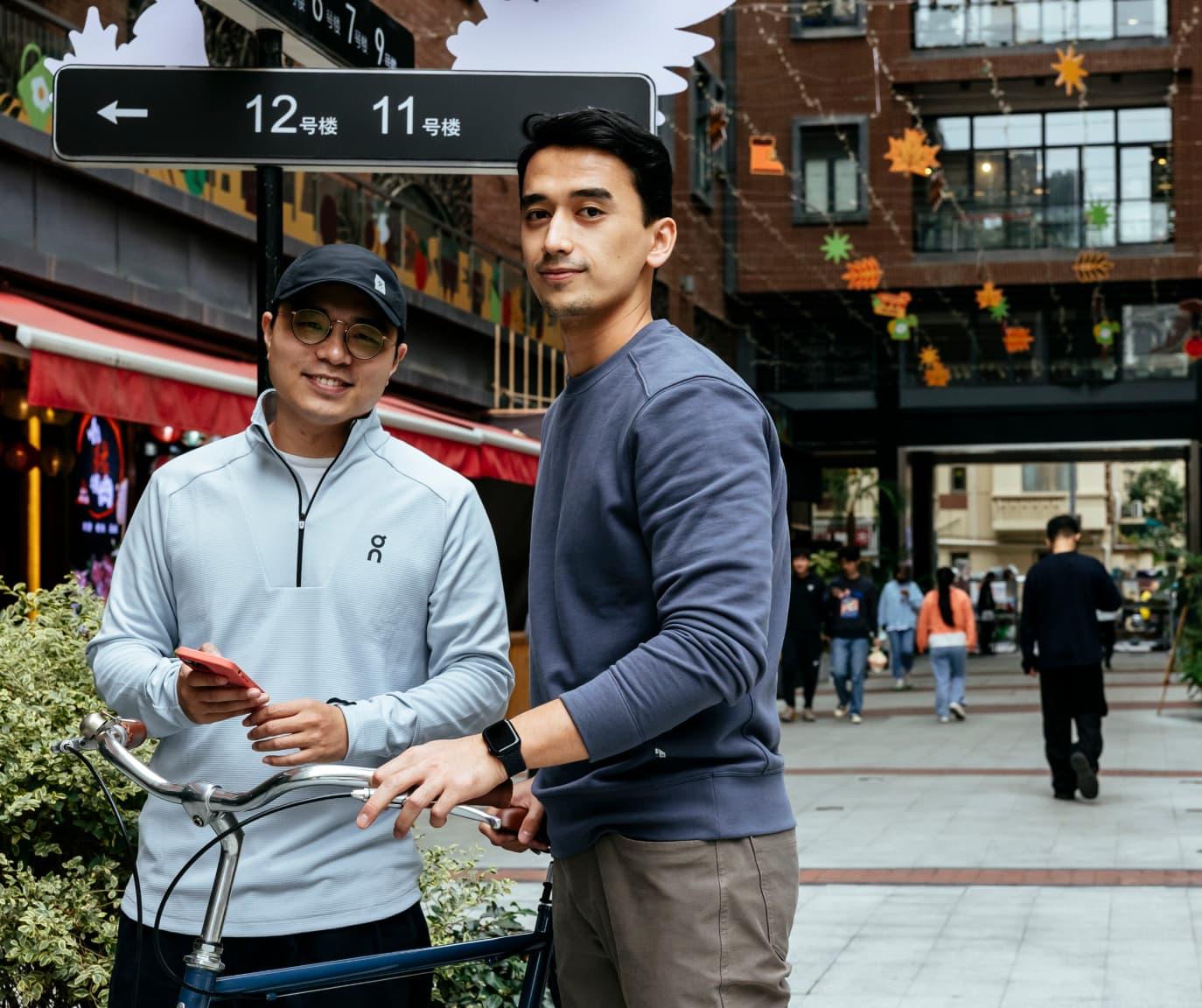 Two men standing on either side of a bicycle, the man on the right has his left hand on the handlebars and the man on the left is holding his phone