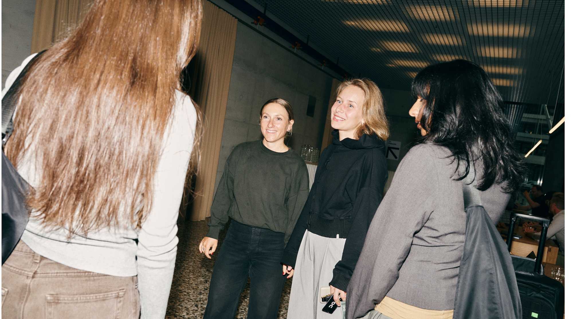 Four women stand and chat in a modern indoor space, smiling and casually dressed.
