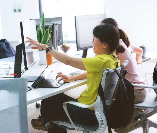 Smiling woman in a yellow shirt points at a computer screen while collaborating with a colleague in a bright office.