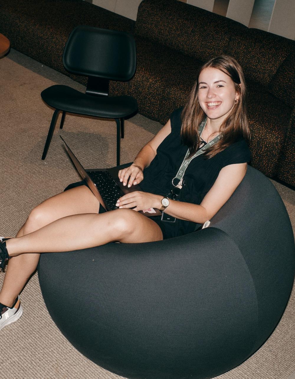 A smiling woman sits in a rounded gray lounge chair using a laptop on her lap while wearing a black outfit and lanyard.