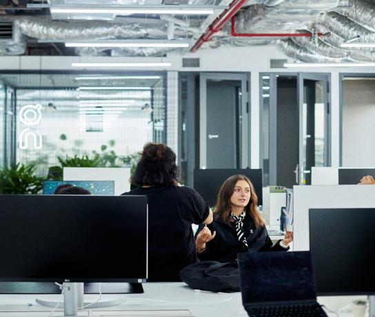 Two people talk across desks in a modern open-plan office with computer monitors and exposed ceiling ductwork.