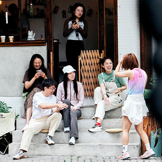 Six casually dressed people sit and stand on concrete steps outside a shop, chatting while one looks at a phone.