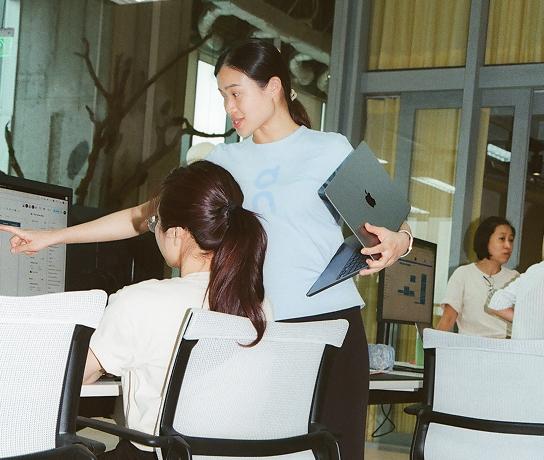 A standing woman holding a laptop points at a computer screen while a seated woman watches in an office.