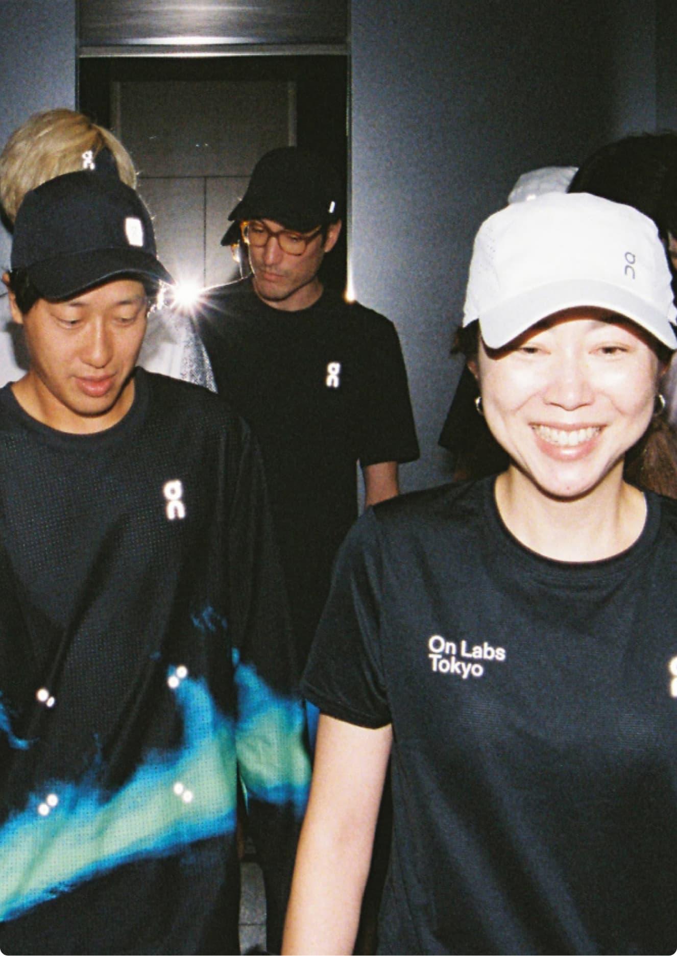 Several people in athletic shirts and caps stand in a mirrored elevator, with a smiling woman wearing a white cap in the front right.