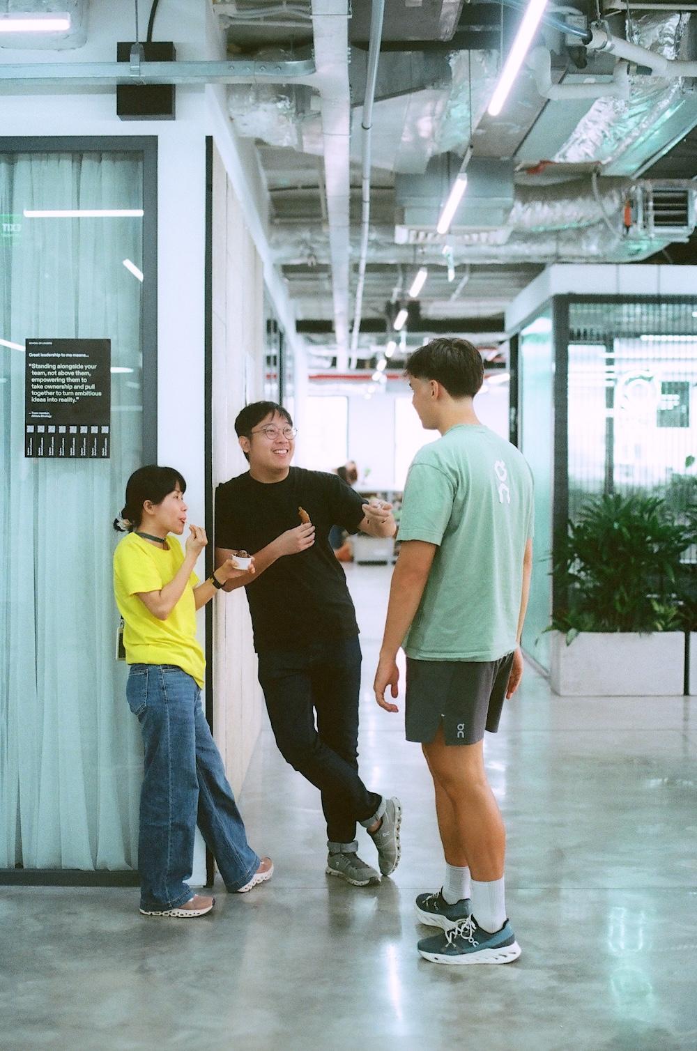 Three colleagues chat casually in a hallway, two eating snacks, with exposed ceiling pipes and plants in a modern office setting.