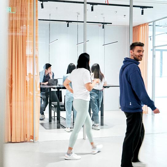 A man in a blue hoodie looks back and smiles as several people walk past a glass-walled meeting room where others stand around a table with a laptop.