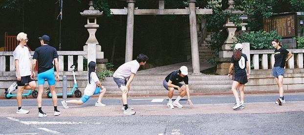 Seven people stretch and talk on a sidewalk in front of stone lanterns and a torii gate.