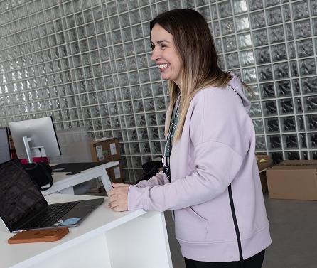 Smiling woman in a light purple hoodie stands at a white counter with an open laptop and phone case, in front of a glass-block wall and stacked boxes.