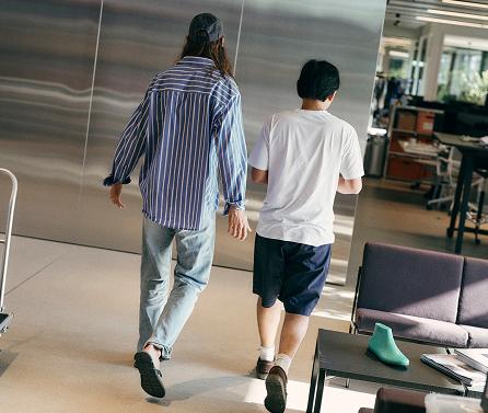Two people are walking away down an indoor space, seen from behind—one wearing a blue striped shirt, jeans and a baseball cap, the other wearing a white T-shirt and dark shorts.
