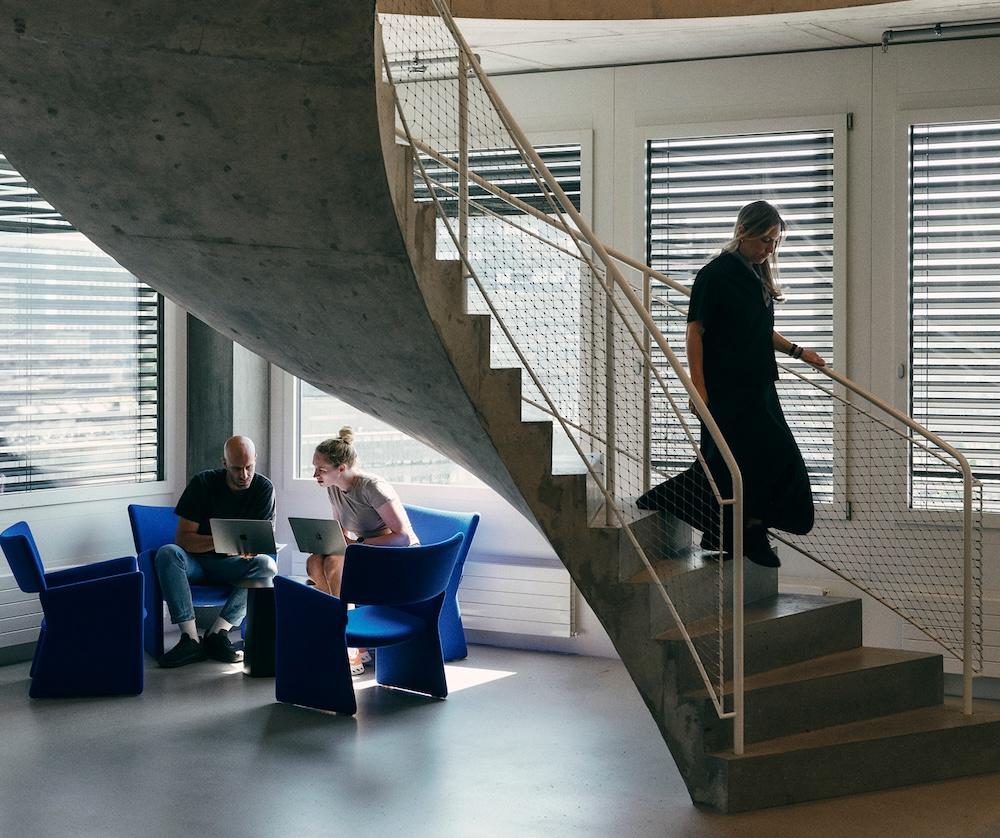 Two people sit in blue chairs under a curved concrete staircase using laptops while a third person walks down the stairs.