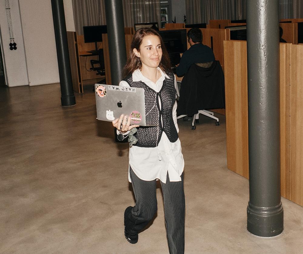 A woman walks through an open-plan office holding a sticker-covered laptop while wearing a white shirt, black knit vest, and pinstriped pants.