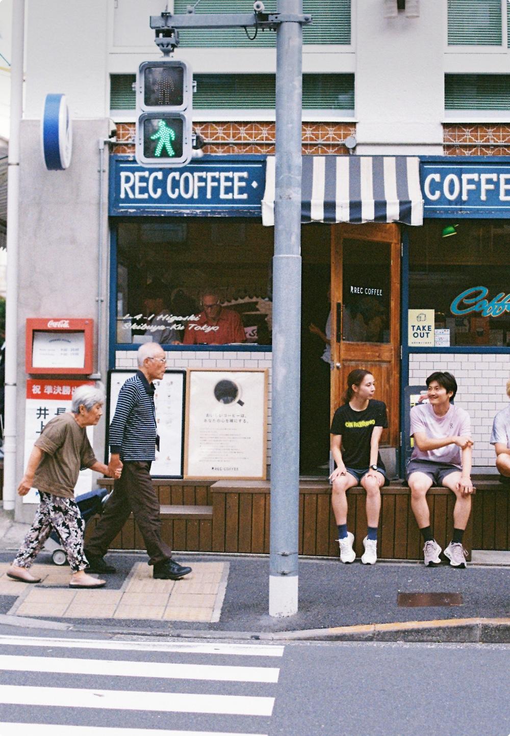 An elderly couple walks past the REC COFFEE storefront as two young people sit on a bench under a striped awning and a green pedestrian signal glows above.