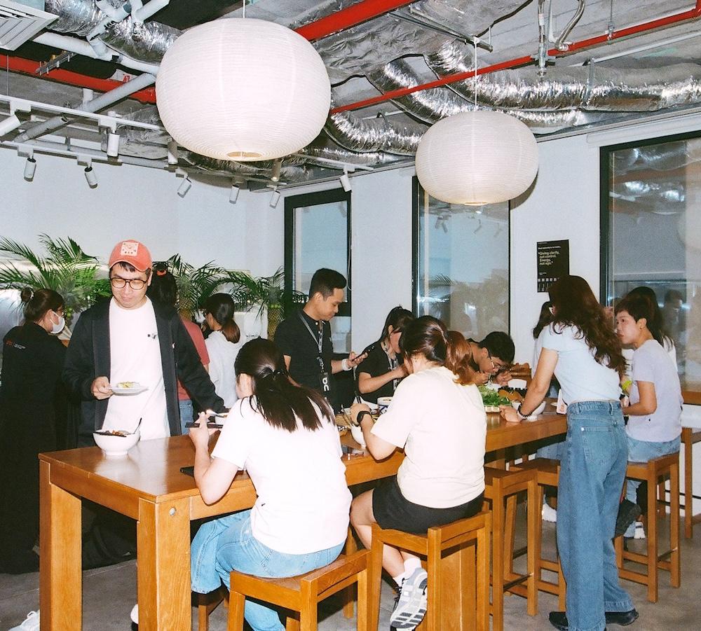Several people sit and stand around a long wooden communal table eating and serving food in a brightly lit dining area with large hanging paper lanterns and exposed ceiling ducts.