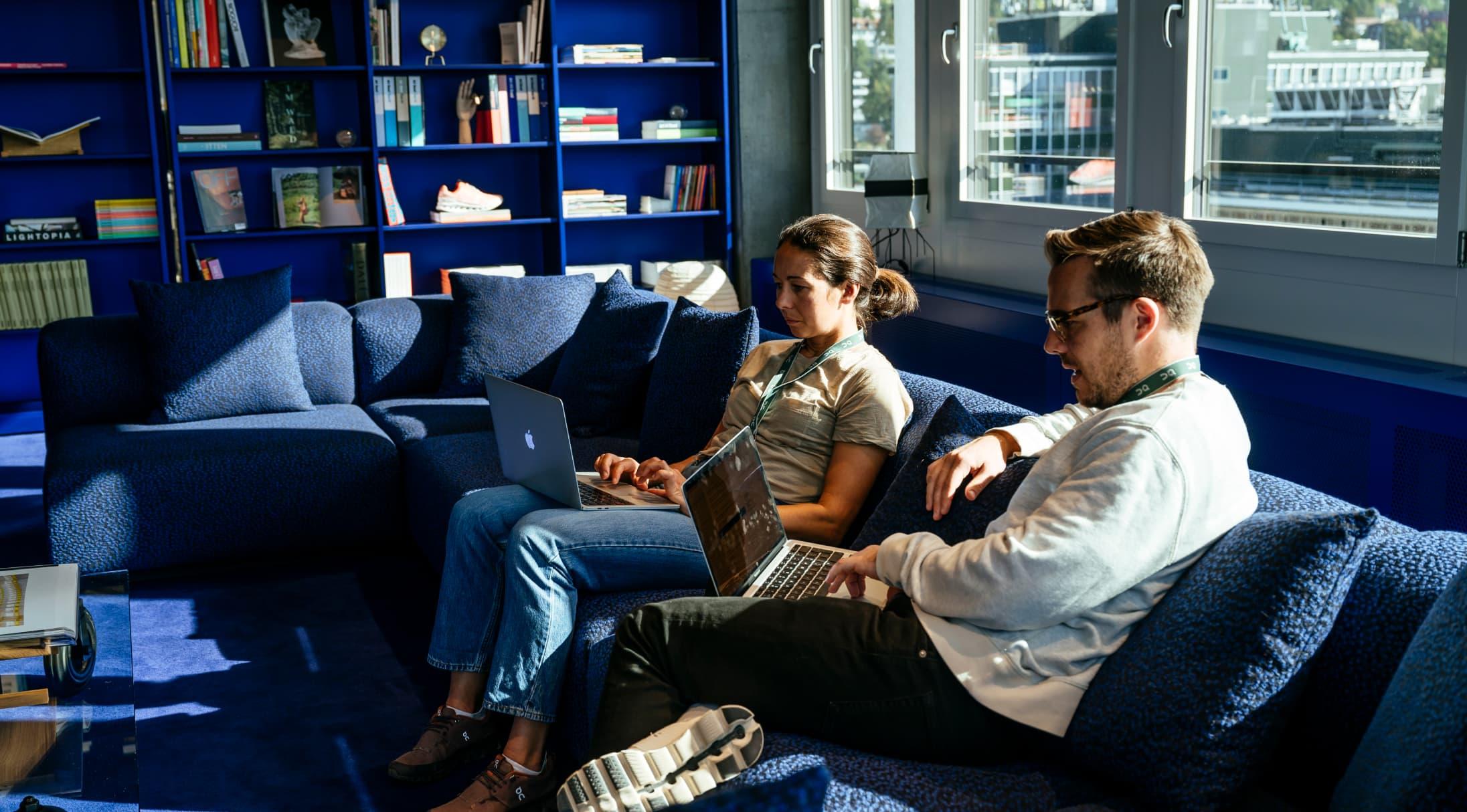 A man and woman are on laptops sitting on a big blue sofa.